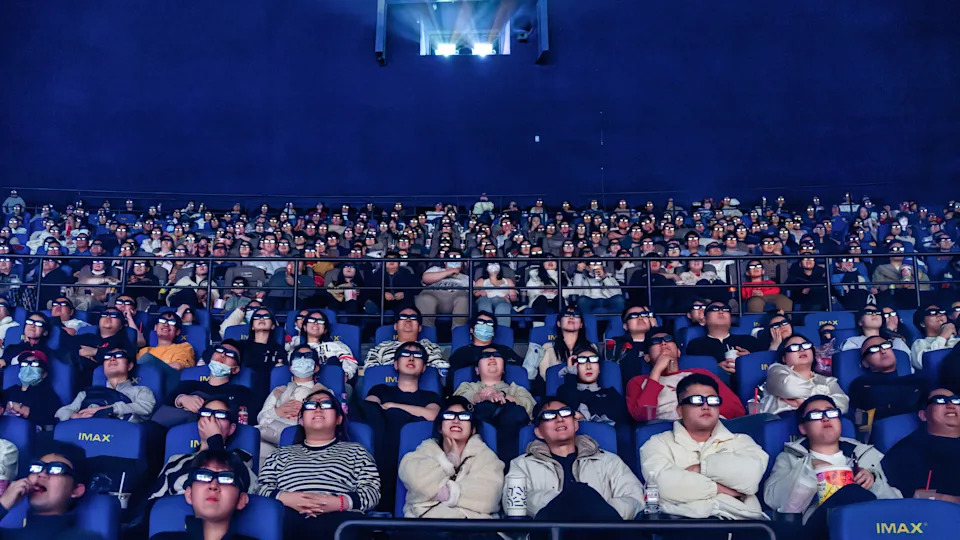 Film pioneers sit in a theater looking at the screen. 